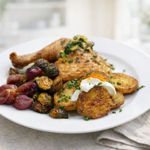 This Is the Best Roasted Chicken Recipe (And It’s Easier Than You Think) 1 roast chicken dinner with roasted potatoes and Brussel sprouts on a white plain in a modern kitchen.